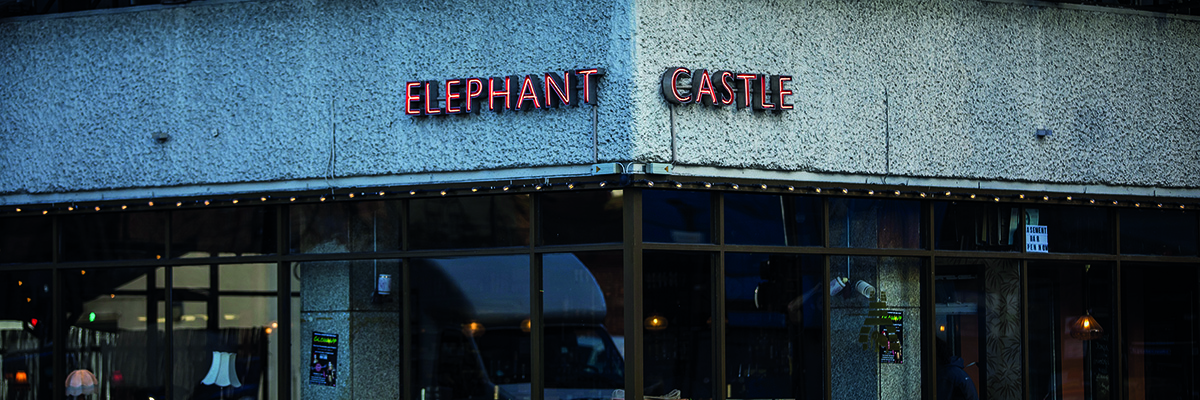 A glass fronted cafe on a street corner. The words 'Elephant Castle' are fixed in red lights onto a strip of grey concrete above the glass frontage. White festoon lighting below.